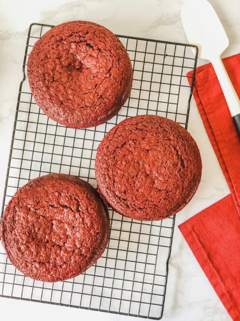 Three unfrosted Red Velvet cake layers cooling on a wire rack.