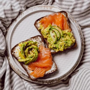 Two slices of toasted bread topped with fresh avocado slices, smoked salmon, and mashed avocado garnished with cucumber and spices, served on a white plate. avocado toast recipe