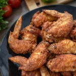 A plate of crispy fried chicken wings on a wooden table with a bowl of ketchup and fresh vegetables in the background.