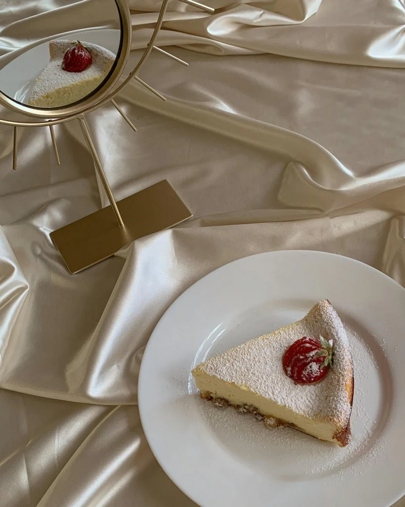 A slice of fluffy Japanese cheesecake on a white plate, dusted with powdered sugar and topped with a fresh strawberry, placed on a silky golden fabric with a decorative mirror reflecting the dessert.