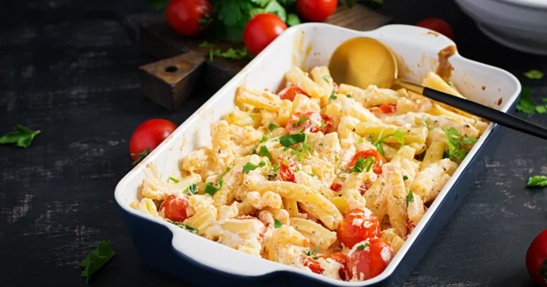 Baked feta pasta in a white oval dish with a gold serving spoon, garnished with fresh parsley and surrounded by cherry tomatoes on a dark rustic background.