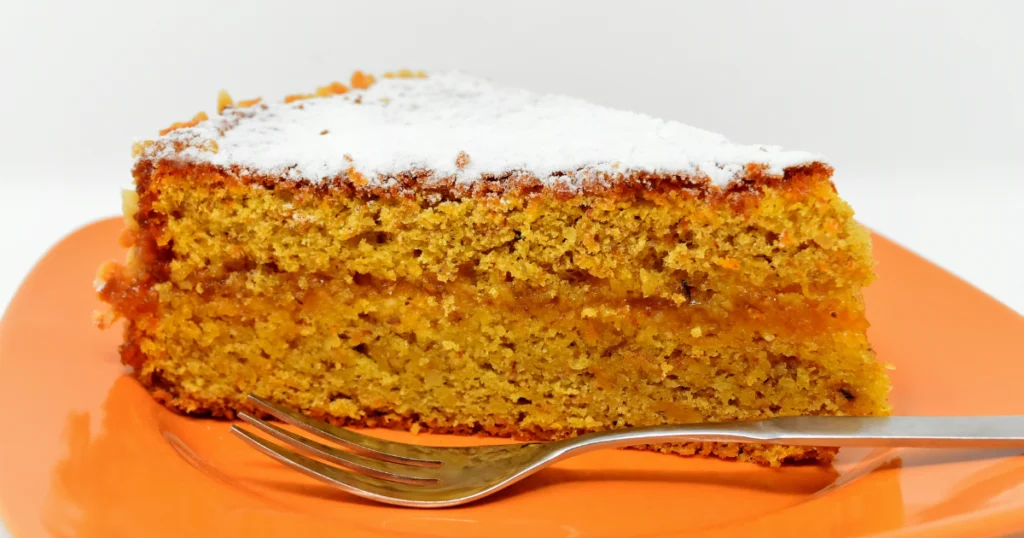 A slice of moist carrot cake with a layer of creamy frosting, dusted with powdered sugar, on an orange plate.