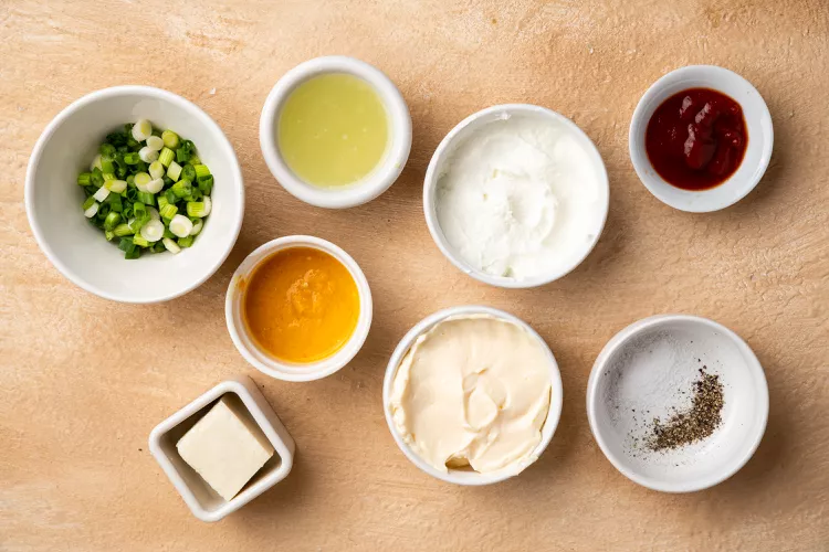 A set of small bowls containing ingredients for aji amarillo sauce, including aji amarillo paste, mayonnaise, yogurt, lime juice, scallions, cheese, ketchup, and seasoning, arranged on a wooden surface.