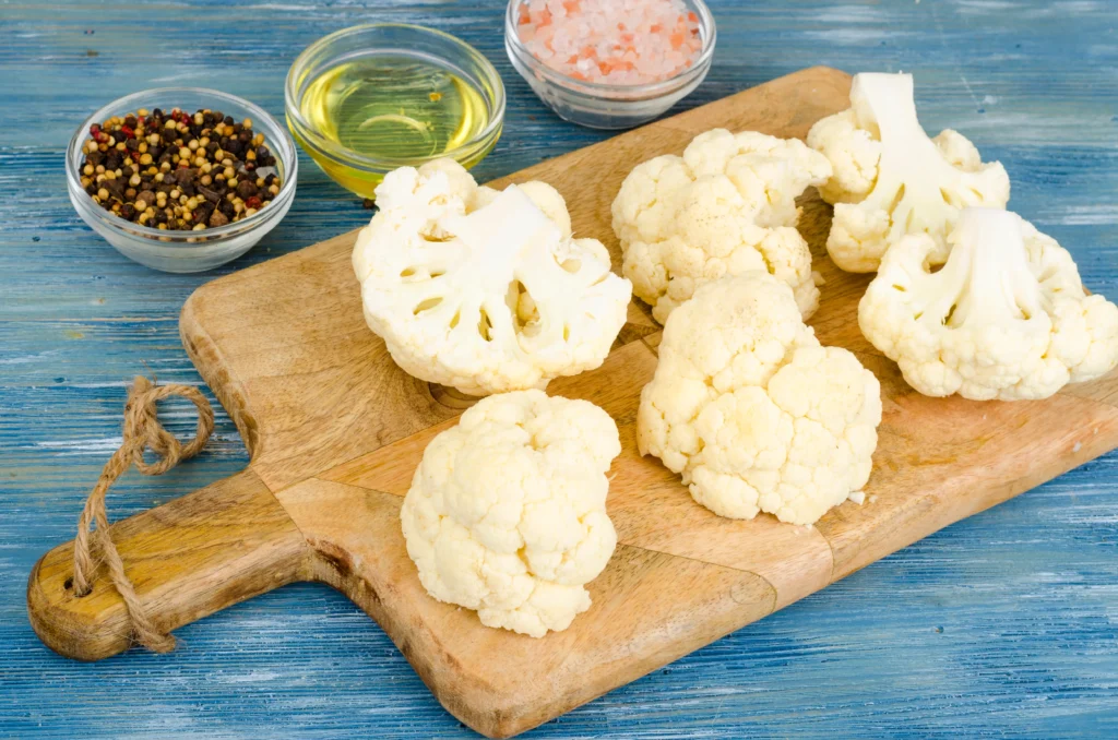 Fresh cauliflower florets on a wooden cutting board with bowls of oil, pink salt, and peppercorns on a rustic blue wooden background.