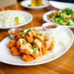A plate of golden, crispy fried chicken bites drizzled with a creamy, spicy bang bang sauce, garnished with fresh parsley and sesame seeds.